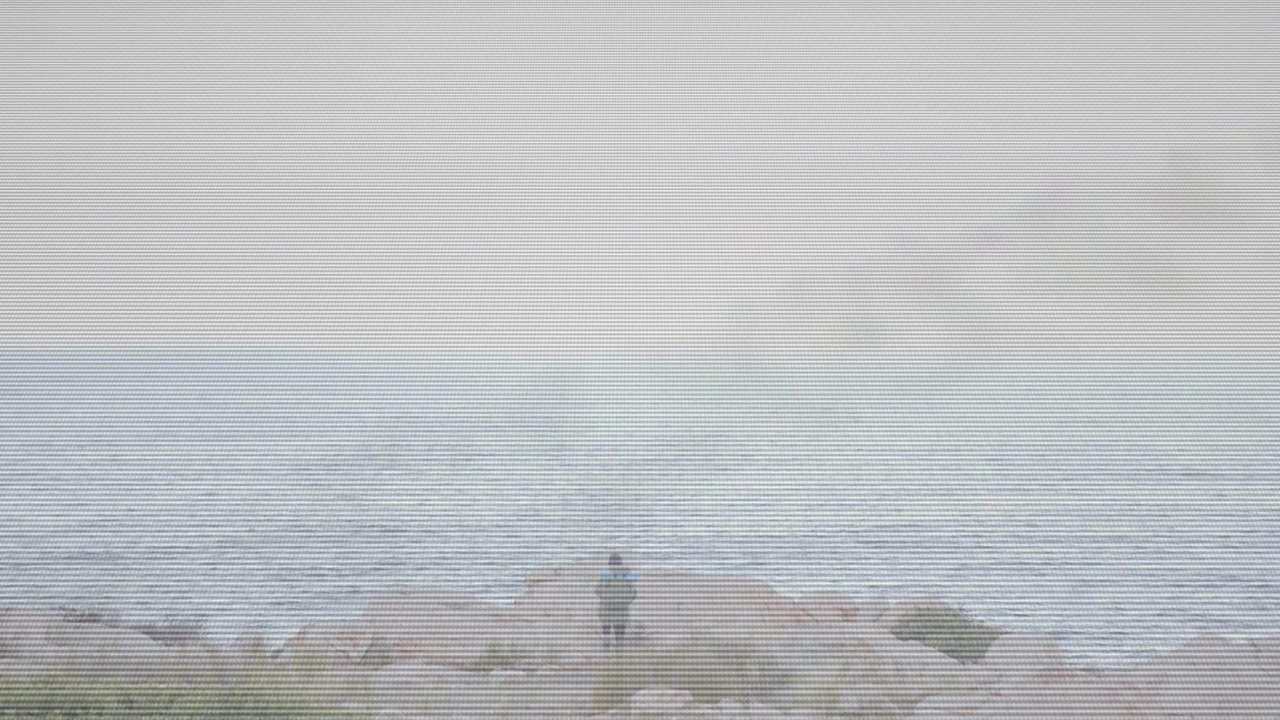 Person appearing lower center initiating measured walking across rocky jetty toward tip to face sea