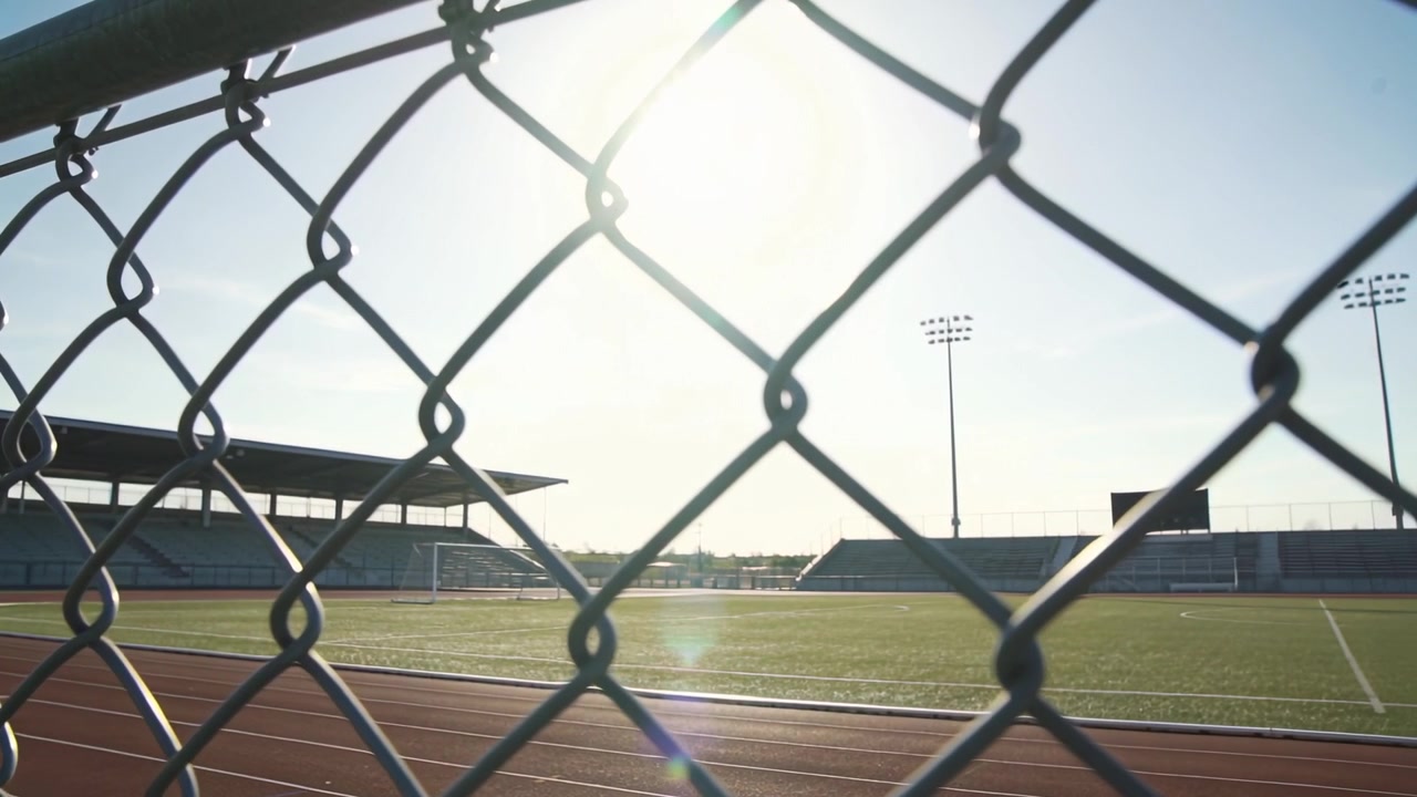 Pulling back camera reacting to sun revealing chainlink fence and track at stadium with lens flare