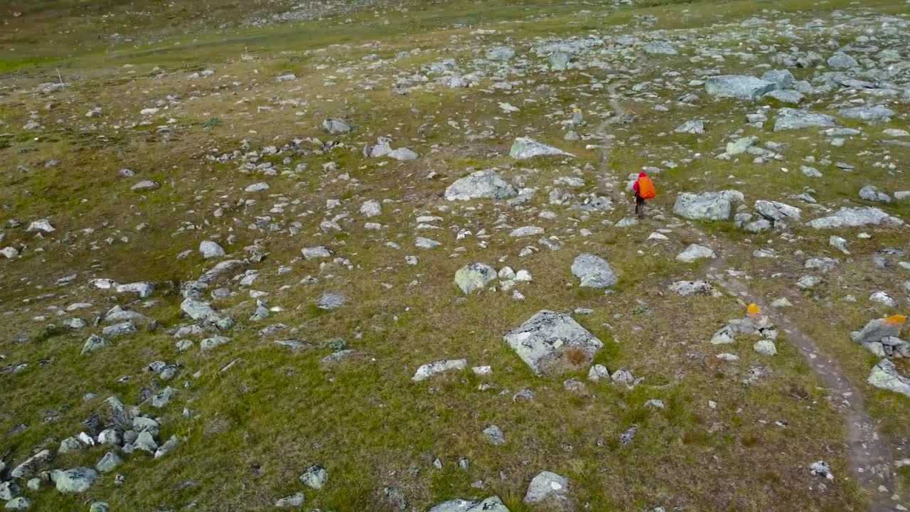 Gorgeous aerial drone footage descending and revealing a backpacker or hiker walking alone on rocky Sweden landscape during a cloudy day while large mountains and hills are visible in the horizon.