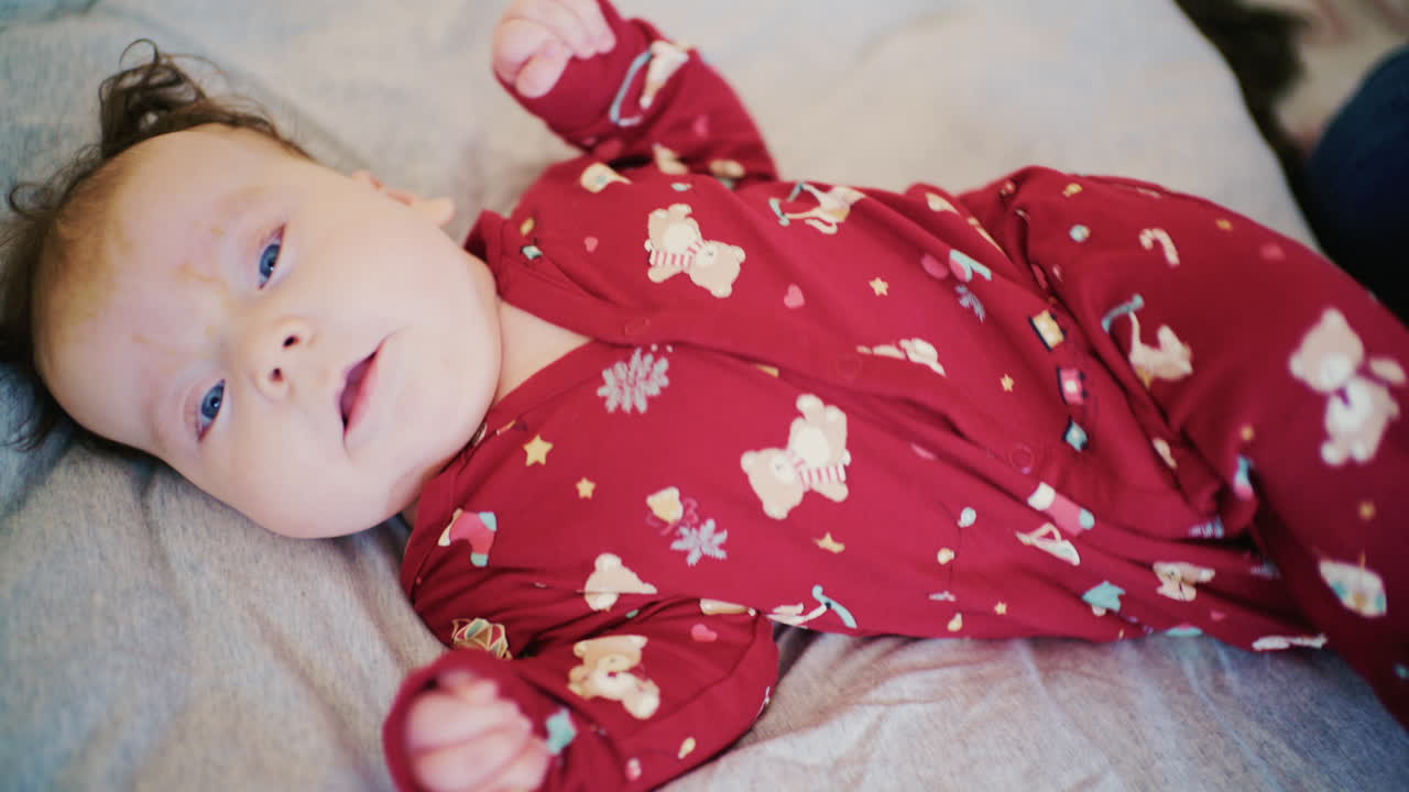 Primo piano di un neonato in tutina rossa sdraiato su un letto mentre la madre esegue esercizi di mobilità.
