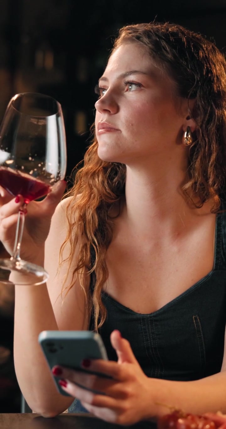 Young Woman Drinking Red Wine While Using Smartphone at Restaurant