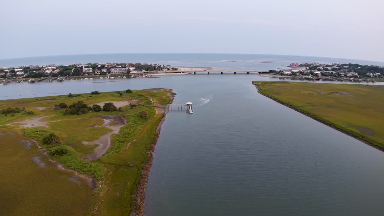 Un drone sorvola dritto un tranquillo canale fluviale fiancheggiato da paludi e zone umide, guidando lo sguardo verso l'orizzonte e il ponte in lontananza