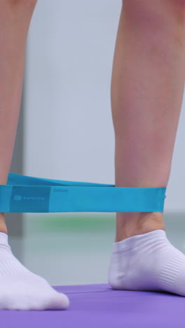 primer plano de una persona atando una banda de resistencia azul alrededor de sus tobillos mientras usa calcetines blancos en una alfombra púrpura una pelota de tenis está suavemente borrosa en el fondo