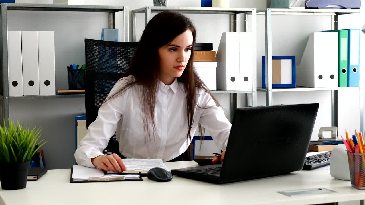 Forretningskvinne ser i dokumenter og jobber på bærbar PC på et moderne kontor