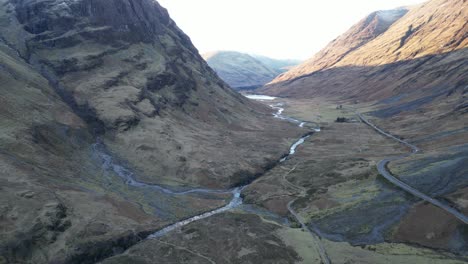 Glencoe-Tal-Mit-Gewundenem-Fluss-Und-Kurvenreicher-Straße-Inmitten-Schroffer-Berge,-Frühes-Morgenlicht,-Luftaufnahme