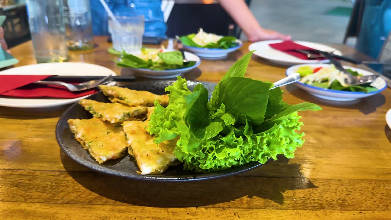 A hand reaches for crispy shrimp pancake garnished with fresh greens on a wooden table in a vibrant Bangkok restaurant