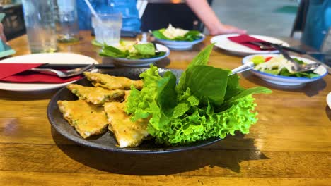 a hand reaches for crispy shrimp pancake garnished with fresh greens on a wooden table in a vibrant bangkok restaurant