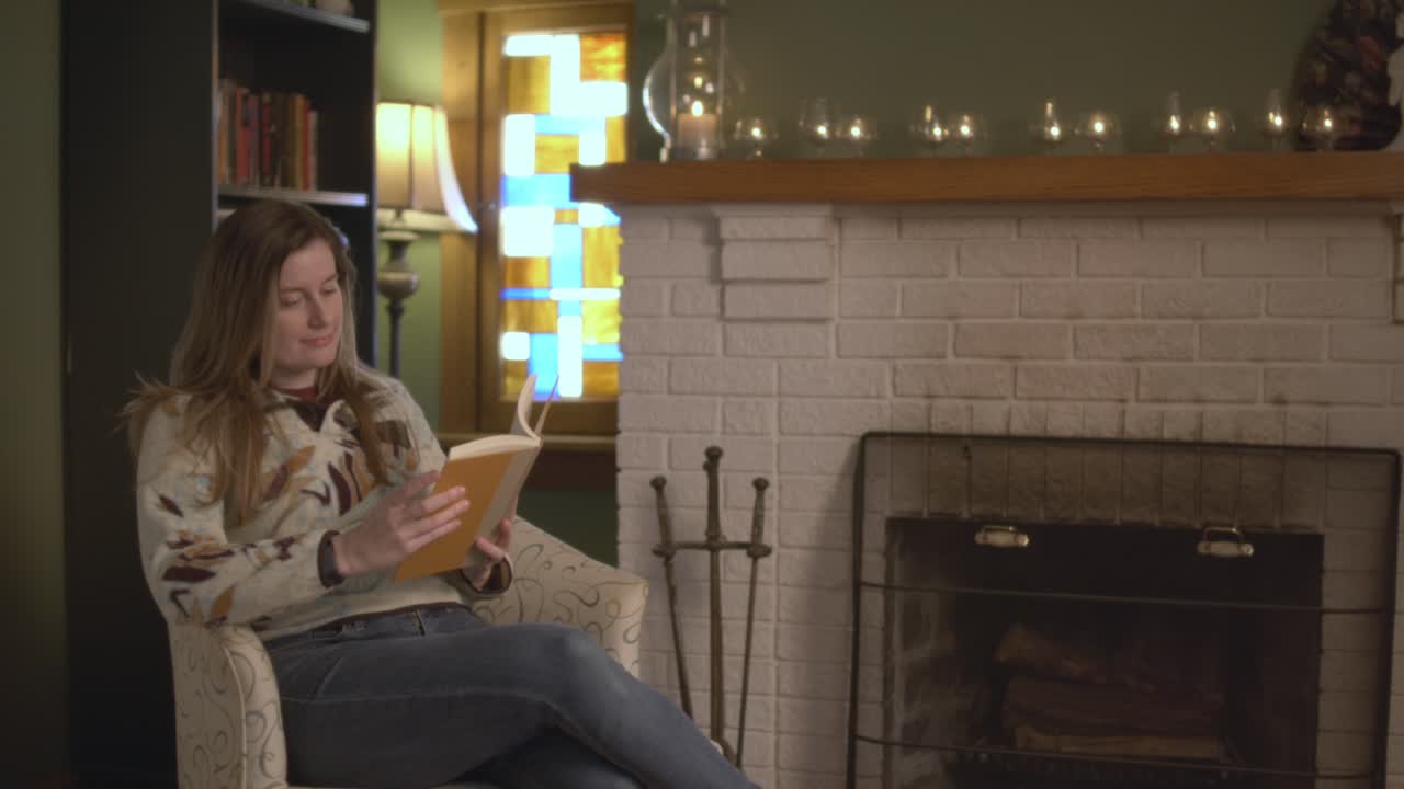 A woman in a sweater walks into a living room, sits down, picks up a book, and reads the book. She sits in a cozy fall location in front of a fireplace. 60 Frames for slow motion