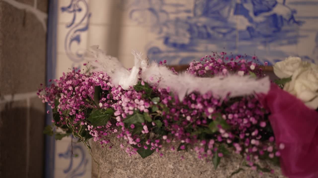 floral arrangement with pink flowers and white feathers on a stone surface in a church, with a religious mural in the background