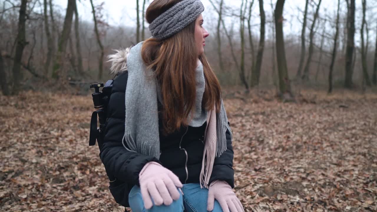 Young female caucasian photographer sitting on stump and looking around her in forest while wearing headband, scarf and pink gloves