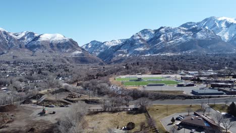 Drone-flying-high-over-small-town-America-with-high-school-football-field,-roads,-and-beautiful-snow-capped-mountains-on-a-sunny-clear-winter-day