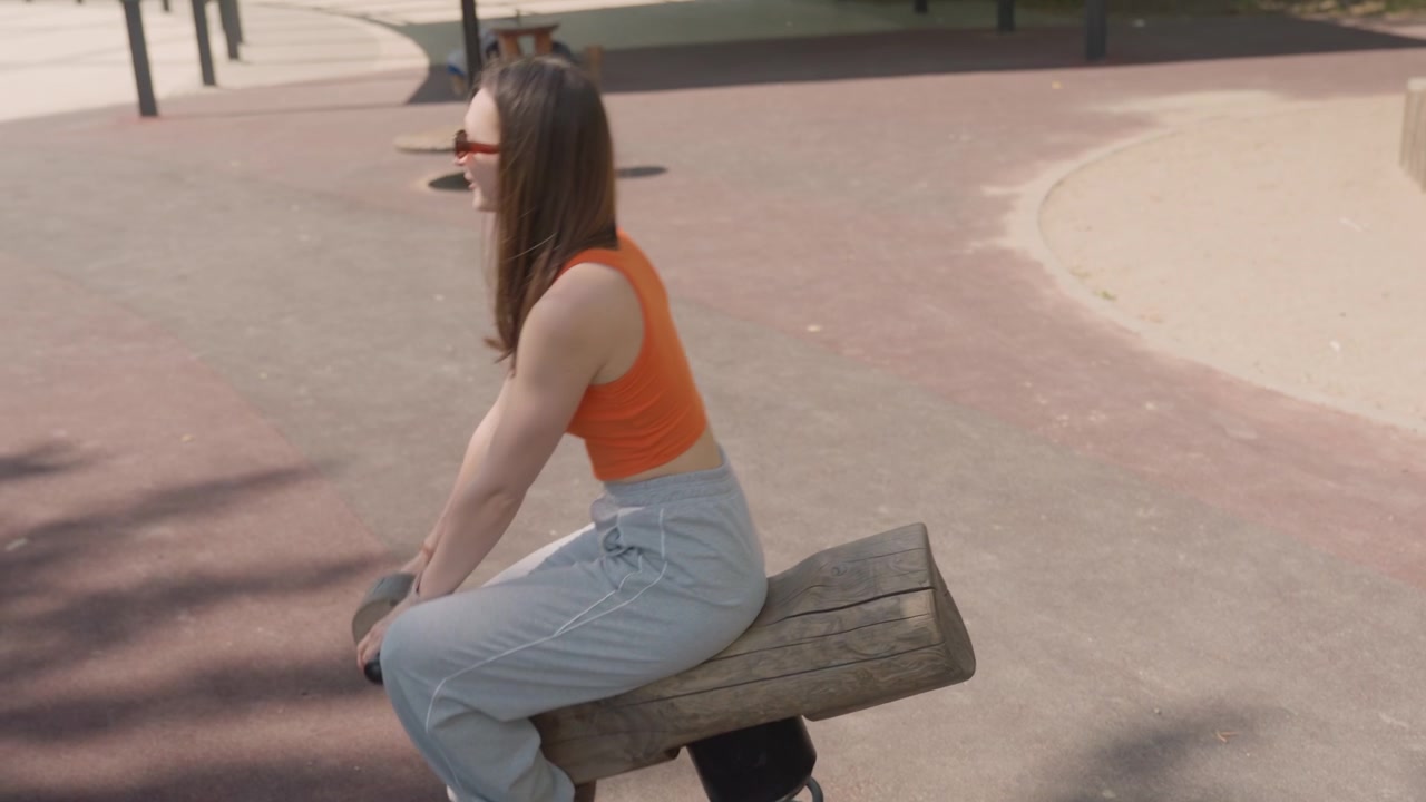 Close up of young woman in orange crop top and gray sweatpants using outdoor gym equipment specifically holding onto handles on wooden spring mounted bench. scene takes place outdoors