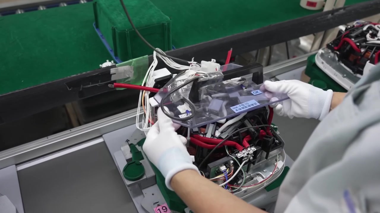 Worker placing protective inspection cover panel onto partially assembled rice cooker on conveyor line