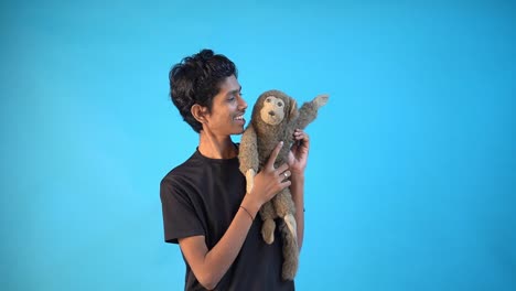 fun young indian boy in basic black t-shirt isolated on blue background studio and playing with toy monkey