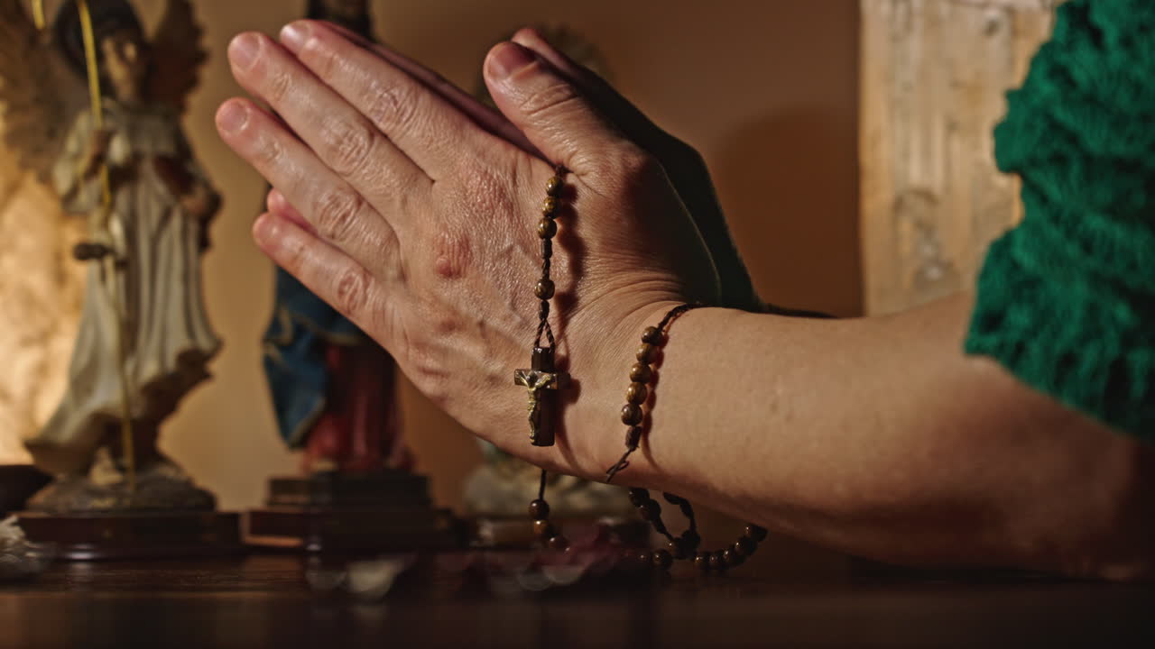 Hands Clasped in Prayer Holding a Rosary with Religious Statues