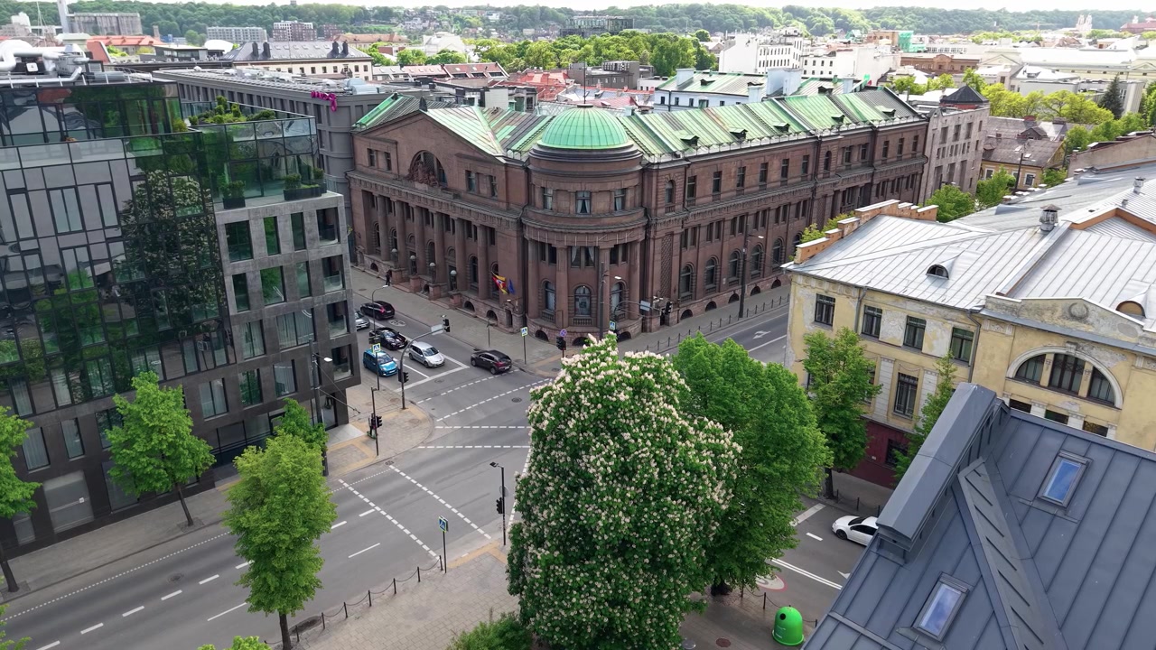 Un video drone cattura un ampio incrocio cittadino incorniciato da un grande edificio storico con cupole verdi, eleganti uffici in vetro, rigogliosi alberi primaverili e una luce diurna vibrante, senza persone visibili