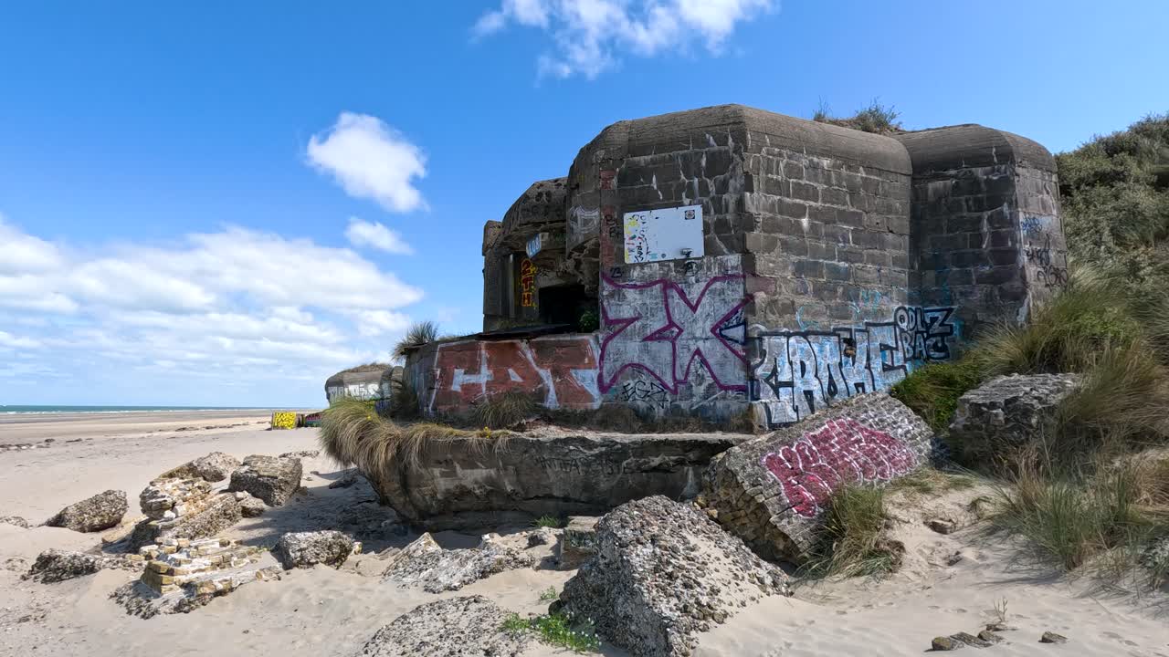 A static wide shot of a historic concrete bunker with colorful graffiti, set on a sandy beach under bright daylight and blue skies in Dunkirk