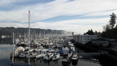 Rows-of-sailboats-lining-the-shore-of-Gig-Harbor-as-Mount-Rainier-is-revealed-above-the-fog,-aerial