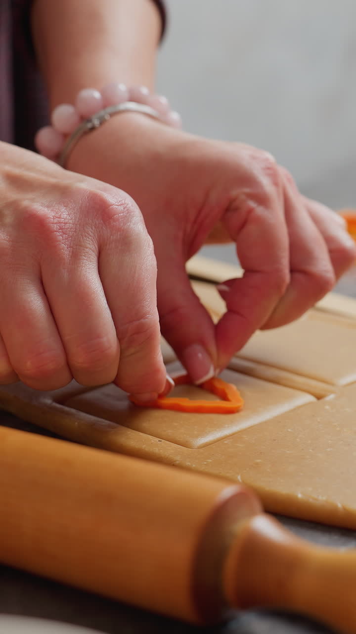 primer plano de las manos de la niña usando un cortador en forma de fresa dentro de una forma cuadrada en la masa, un rodillo de madera descansa al lado del espacio de trabajo, con otros cortadores de galletas naranjas esparcidos en el fondo