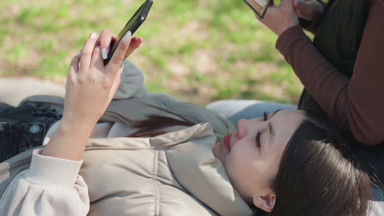 아시아 여성이 풀밭에 누워 문자를 보내고, 옆에서는 백인 친구가 책을 읽고 있는 따뜻한 봄날의 공원 풍경. 햇빛 아래 재킷을 입고 편안한 자세를 취하고 있는 모습에서 우정이 느껴지는 자연스러운 순간, 스크롤과 책장 넘기는 모습