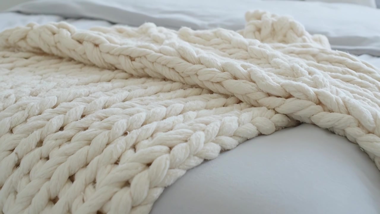 Telecamera a bassa angolazione che si sposta rivelando la trama di una spessa coperta lavorata a maglia color crema sul letto, cuscino visibile