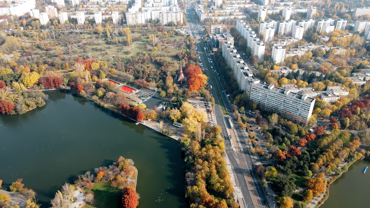 A road over the the lake with moving cars in the Titan park, multiple greenery and residential buildings. View from the drone, Bucharest, Romania