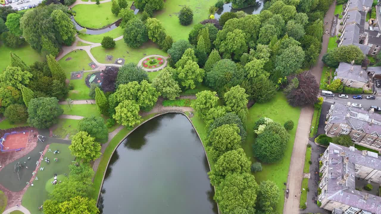 Drone footage glides above a lush town park in Buxton, England, revealing a pond, walking paths, and residential buildings under soft daylight