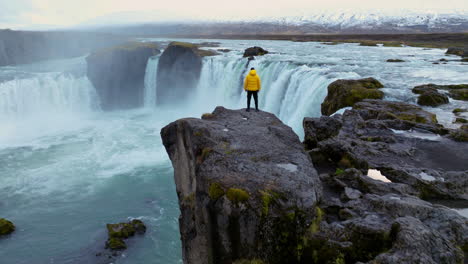 man standing on a cliff next to godafoss waterfall in iceland - aerial pullback