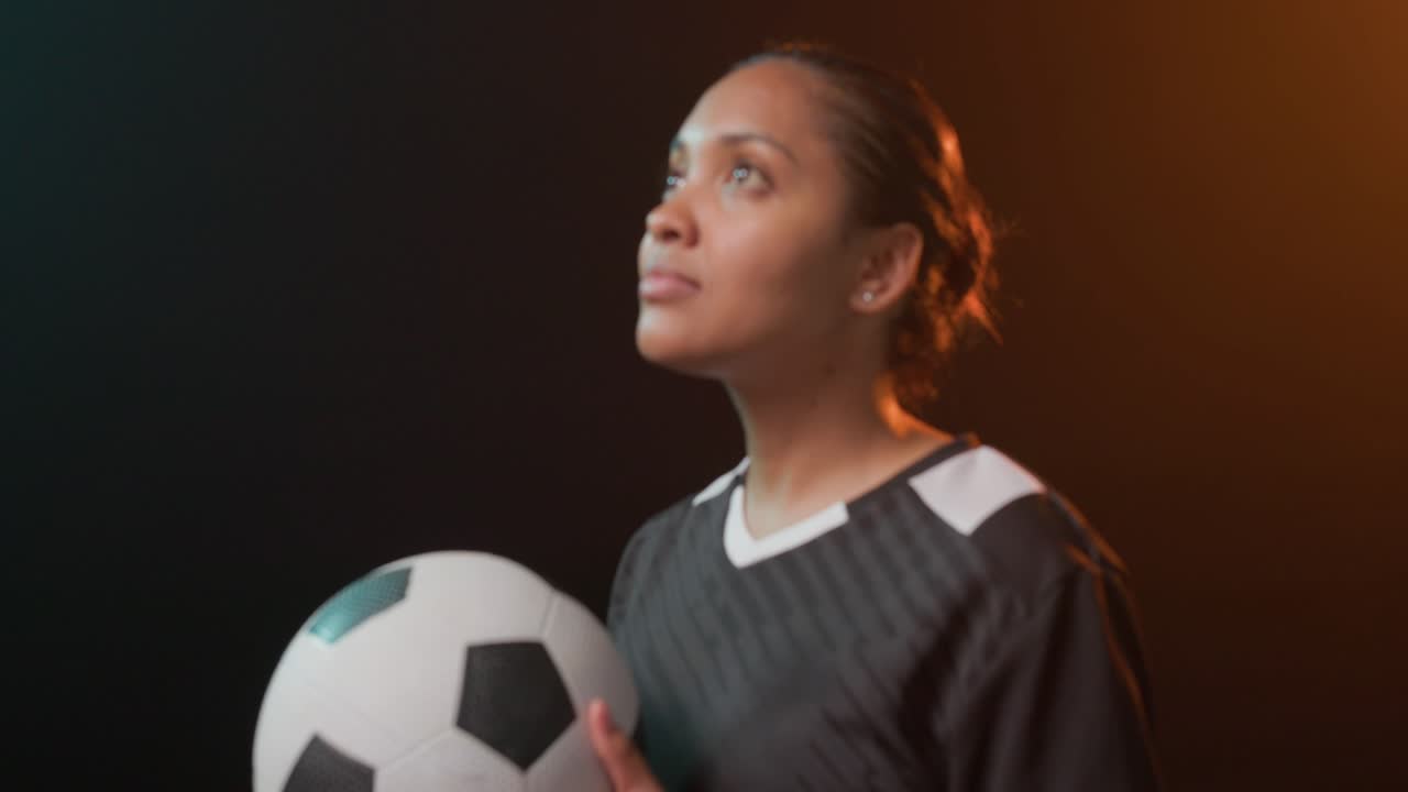 Donna con pallone da calcio, maglia nera, in studio, contorno verde acqua-arancione, primo piano della telecamera, che guarda in alto.