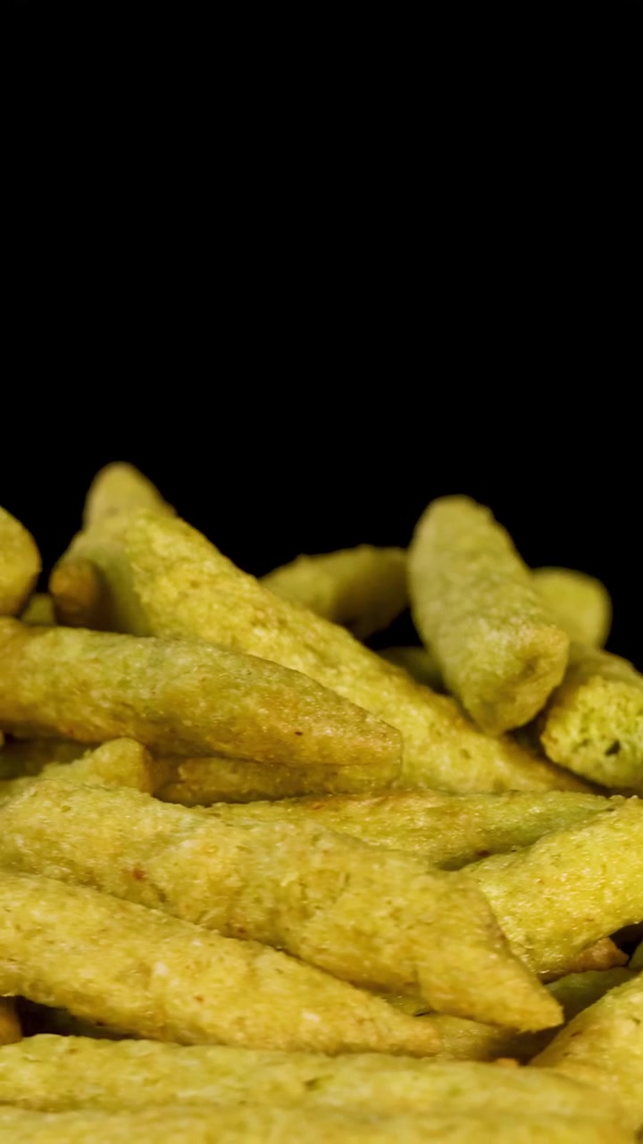 Vista macro rotante di bastoncini snack di piselli verdi soffiati su sfondo nero, con illuminazione da studio uniforme.