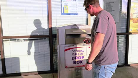 man votes in election by dropping mail-in ballot letter in slot at voting booth with offical ballot drop box sign for democratic government campaign in presidential race