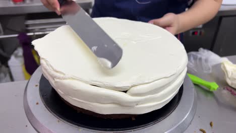 pastry chef using spatula to frost a smooth cake with buttercream meringue frosting the whole cake while spinning the revolving cake turntable stand, close up shot in commercial bakery setting