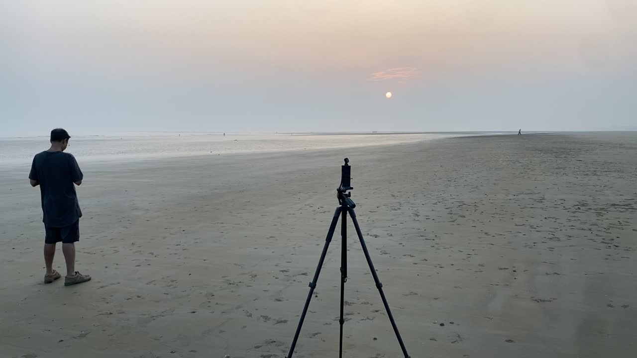 POV of an Indian man standing with phone and camera stand tripod in a clear pleasant empty beach with beautiful sunset.