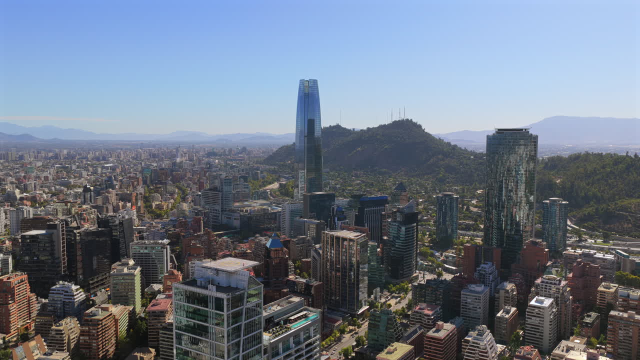 Veduta aerea di Santiago che mostra moderne torri di vetro, densi edifici urbani e le colline vicine sotto la chiara luce del giorno.