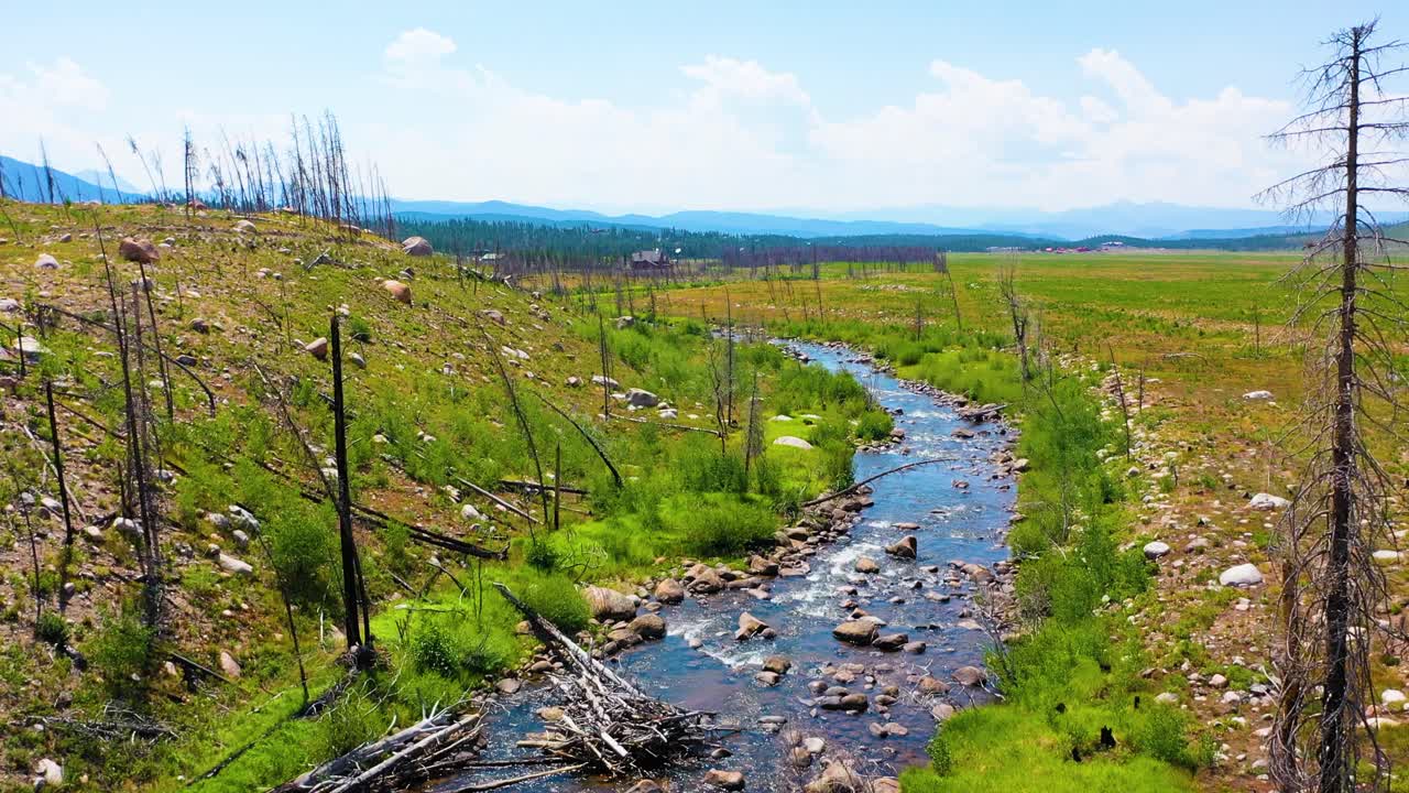Framåtgående flygfilm i Rocky Mountain National Park nära Grand Lake, Colorado, som följer en slingrande bäck över öppen ängsterräng med spridda stenblock, förkolnade träd och bergsutsikt.