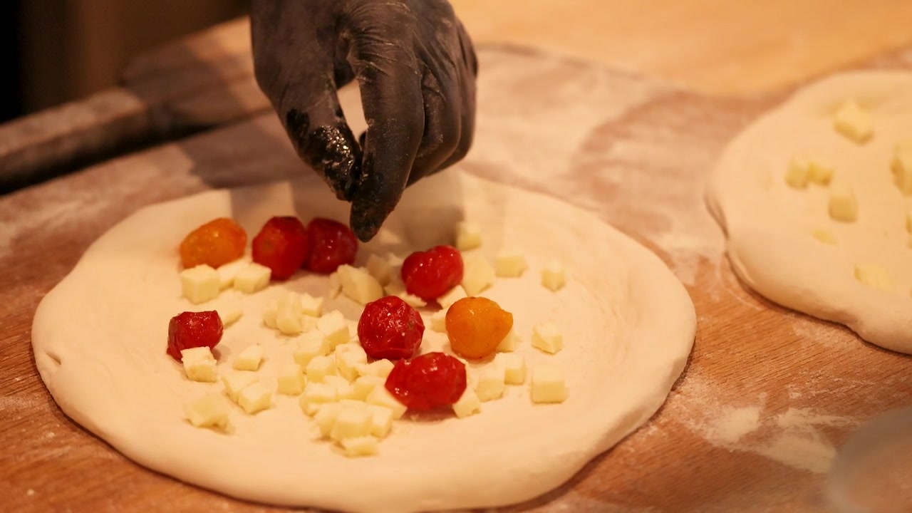 Primo piano di mani che guarniscono impasto crudo per pizza con mozzarella e pomodorini ciliegino
