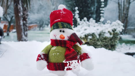 a snowman sof toy is sitting on a table in a garden as snow falls around him