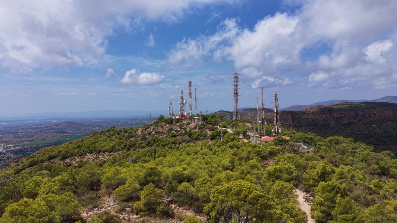Multiple telecommunication towers and antenna masts atop a green Mediterranean hilltop under a blue sky with clouds, overlooking a vast valley landscape