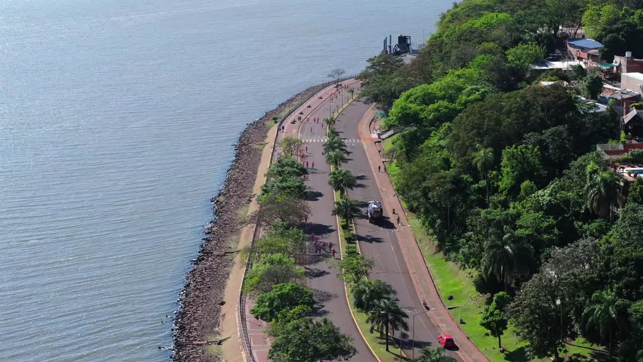 Drone che effettua una panoramica da sinistra a destra sul lungomare di Posadas, mostrando dei corridori su una strada costiera, alberi, lungomare e il fiume Paraná