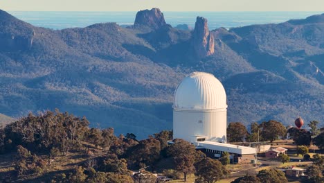 aerial drone footage orbits a large white observatory dome at sunset, revealing rugged mountain ranges and forested landscape under warm, natural lighting