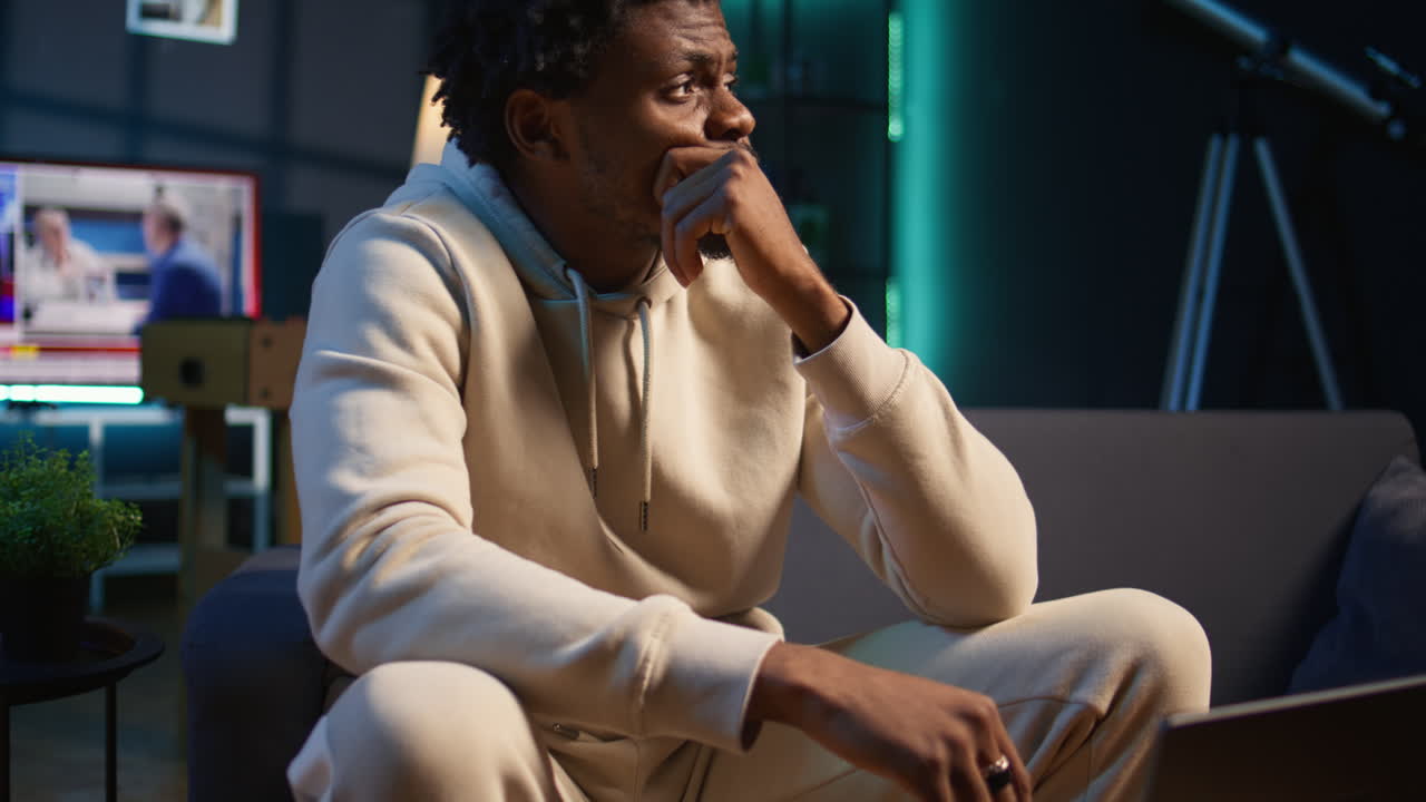 African american male remote worker sitting at home on couch late at night, overthinking about problems. Young man struggling to finish work project before deadline, finding solutions. Camera A.