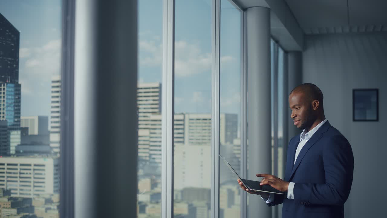 Thoughtful Black Businessman in a Tailored Suit Using Laptop while Standing in Office Near Window on Big City. Successful Corporate Top Manager Doing Data Analysis for e-Commerce Startup
