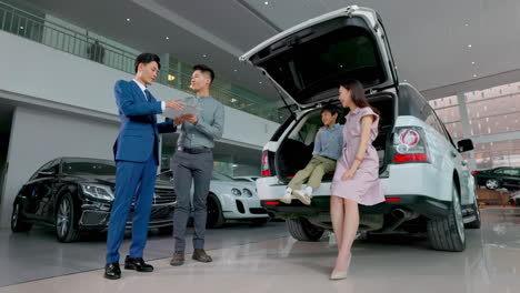 happy couple with attractive auto dealer in a suit presenting new car model to his client, shake hands with salesman and take key to new car