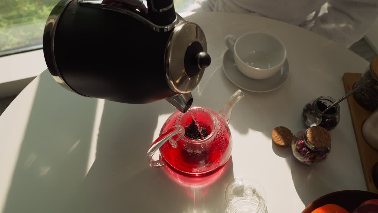 Process of crafting wholesome beverage. Half-full teapot of brewed tea continues filling with water from kettle. Man makes natural fruit tea