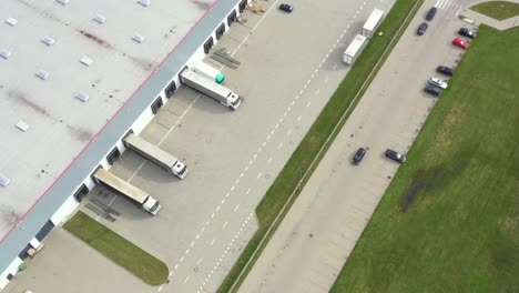 aerial following shot of semi-trailer trucks travelling along a warehouse on the parking lot in the logistics park with loading hub