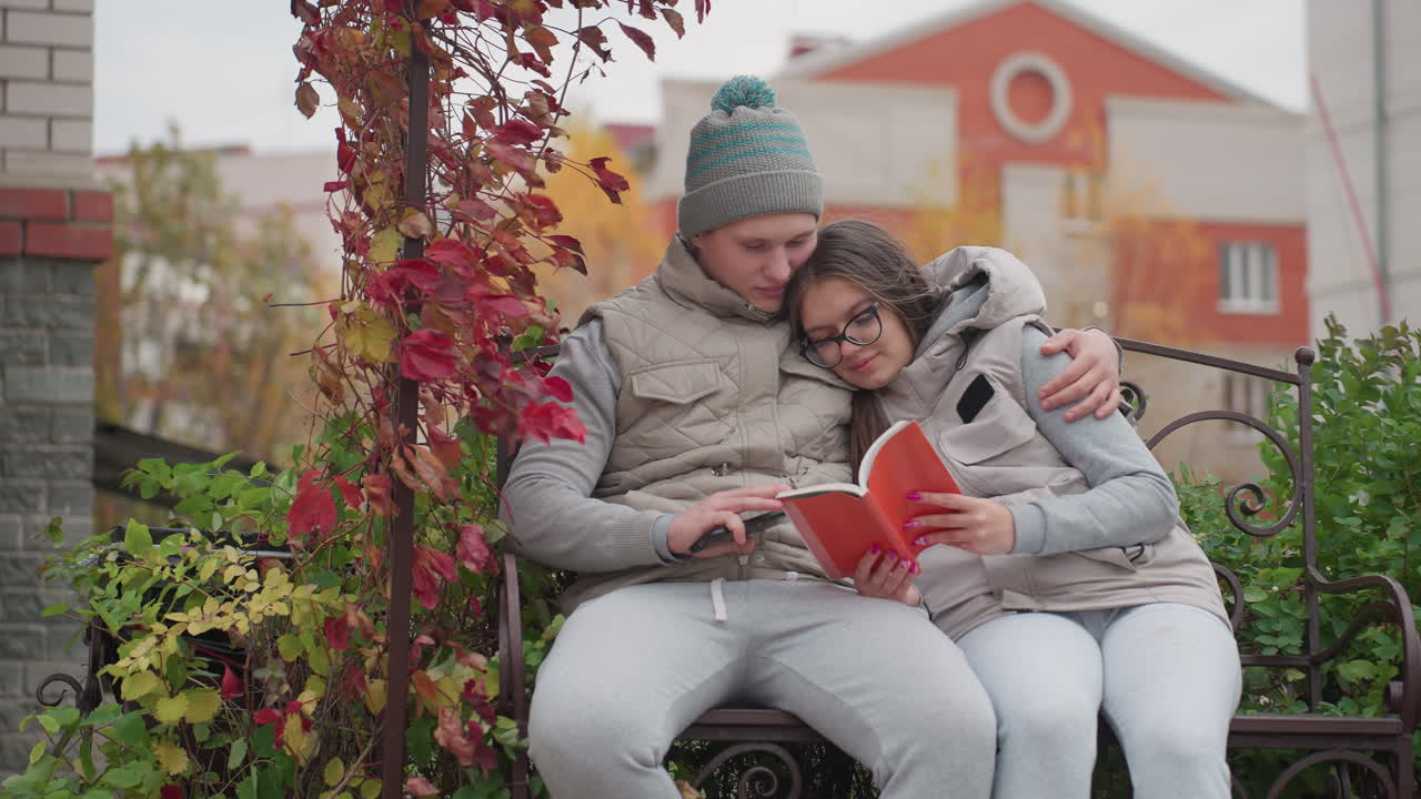 Man and wife seated outdoor park bench enjoying peaceful moment as wife rests on husband while both read book together with man hand on book holding phone gently as breeze sways