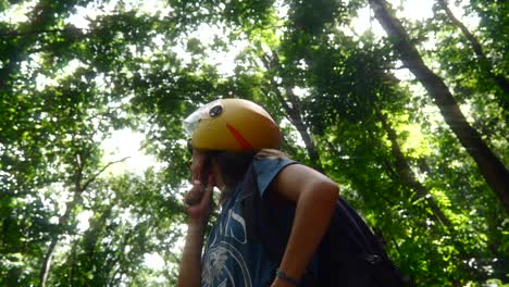 Woman-adjusts-helmet-strap-as-she-looks-around-at-dark-green-tropical-jungle-on-sunny-day