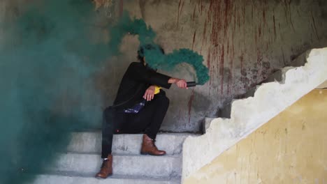 hombre de raza mixta sosteniendo una granada de humo con humo verde sentado en los escalones de un almacén sucio