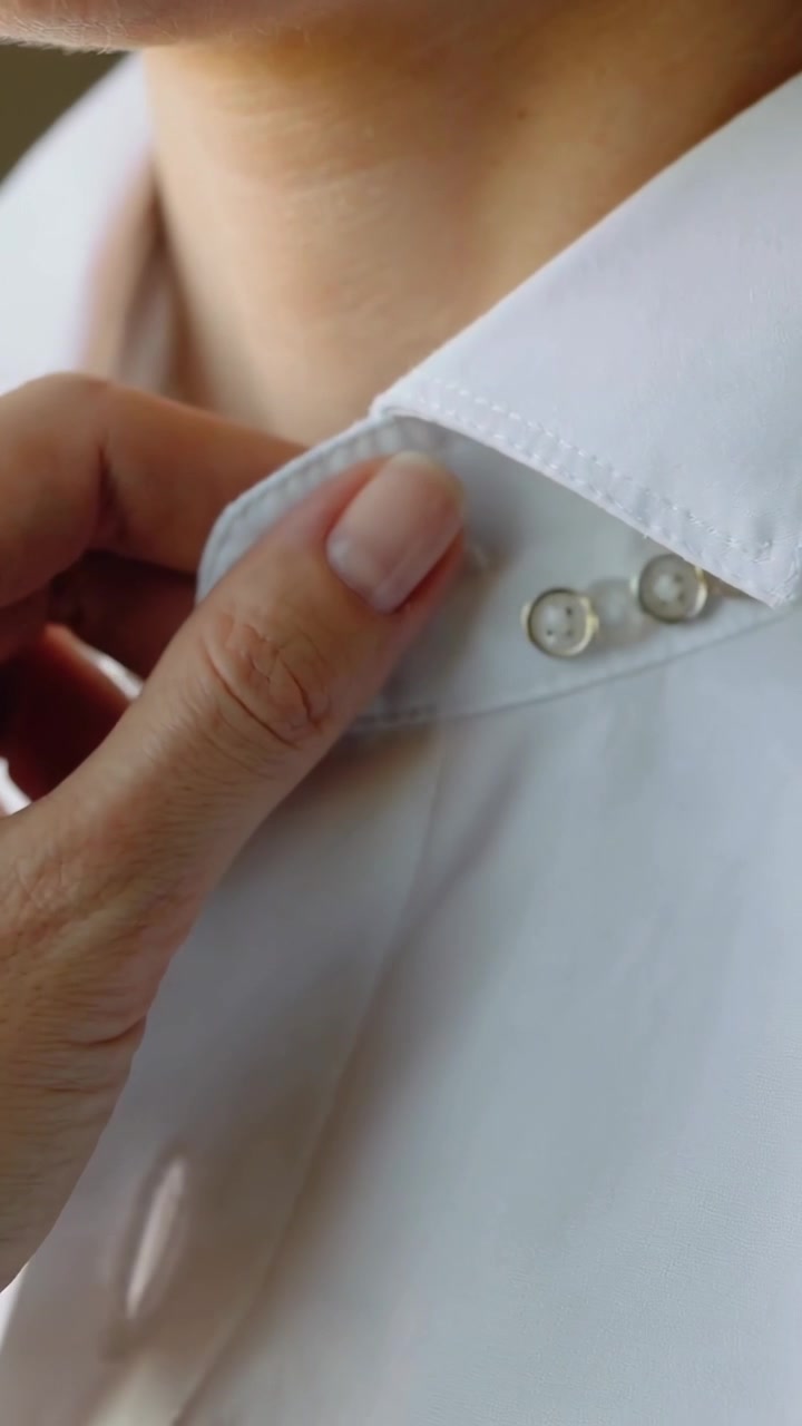 Video verticale: Mentre si prepara, una mano curata solleva il colletto di una camicia bianca a casa, mostrando una cucitura rossa.