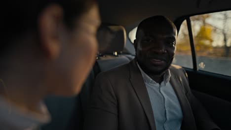 Over-the-shoulder-a-man-with-a-beard-and-Black-skin-in-a-brown-jacket-communicates-with-his-colleague,-a-brunette-girl-with-round-glasses-during-his-trip-to-a-modern-salon-in-a-car-outside-the-city